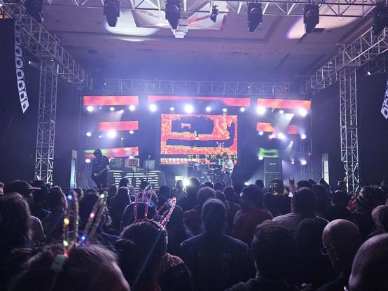 A three-piece band performs to a large crowd in front of a screen showing footage from the Amiga game Lemmings.