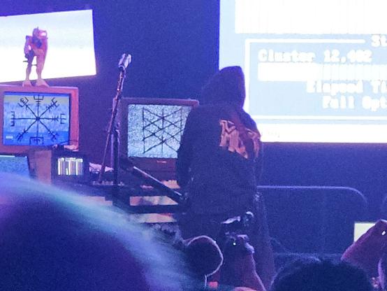 A person in a hoodie works the keyboard of a Commodore commputer on a stage, with two CRT monitors in front of them.