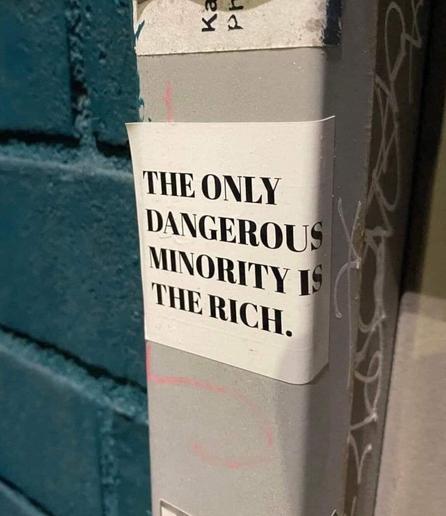 A bold black-and-white sticker with capitalized text reading, "THE ONLY DANGEROUS MINORITY IS THE RICH." The sticker is placed on a metal pole covered in graffiti and scratches, positioned next to a textured blue brick wall. The statement is a striking political message critiquing wealth inequality, blending seamlessly into the urban environment.