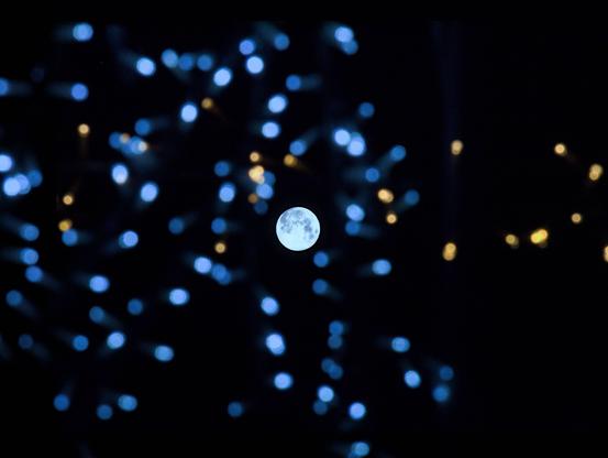 L'image montre la lune visible à travers une constellation de lumières colorées. Les lumières sont floues et apparaissent comme un bokeh jaune et bleu sur le ciel nocturne sombre. La lune est mise au point, bien en évidence au centre de l'image.