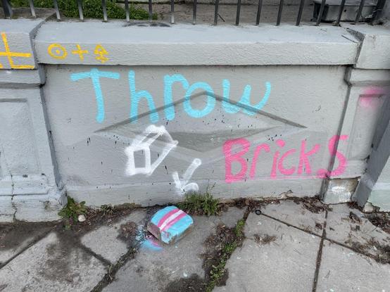 Graffiti in blue, white, and pink, that says "Throw Bricks" with an arrow pointing to a brick that has been painted like a trans flag.