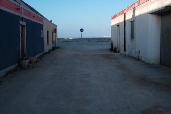 Photo of a small alley, with old, colored houses on both houses and a traffic sign pointing towards the left at the end.