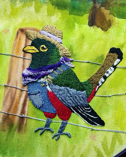 Un pájaro bordado con un sombrero de paja y un chaleco de mezclilla se encuentra sobre una valla de alambre de púas, contra un fondo verde con árboles borrosos. El pájaro es colorido, con tonos de verde, rojo, azul y negro.