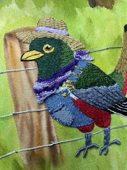 Un pájaro bordado con un sombrero de paja, un mono de mezclilla y un pañuelo morado se encuentra en una valla contra un fondo verde. El pájaro tiene una cabeza verde, pico amarillo y detalles rojos en su cuerpo.
