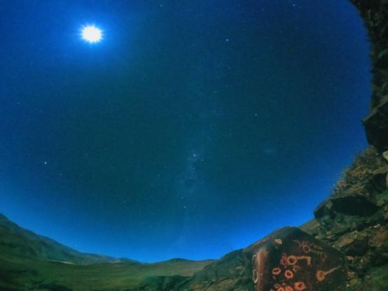 Ciel bleu foncé sphérique avec un point lumineux, le soleil, quelques étoiles et constellations qui compose l'essentiel de l'image... Autour des massifs rocheux verdâtres avec au premier plan des rochers dans les mêmes tons et d'autres marron-rougeatres sur lesquels il y a des formes géométriques comme gravées, orangées