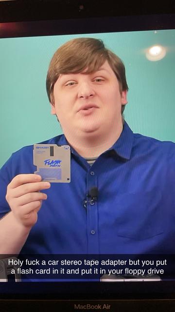 A person is holding a floppy disk labeled "FLASH PATH" while speaking excitedly. They are wearing a blue shirt and are set against a light green background. The image includes text overlay expressing surprise about a device that resembles a car stereo tape adapter.
