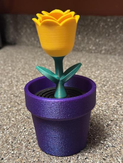 A 3D printed flower in a pot. The flower is a Rose. The flower head is yellow, the stem is green, and the pot is purple.
