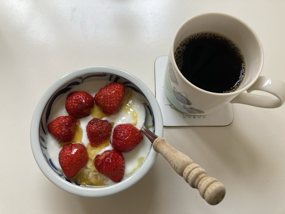 A bowl of porridge with yogurt, honey, and seven small strawberries on top, with a mug of black coffee.