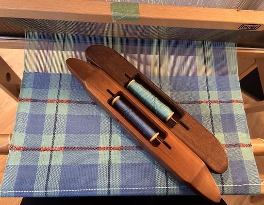 Two handmade wooden boat shuttles with different colours of blue cotton yarn sit obliquely across woven cloth on a hand loom