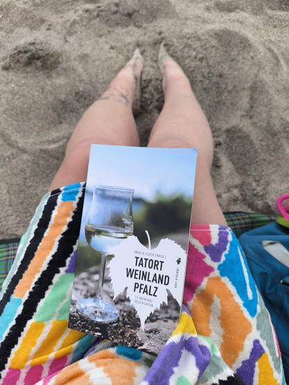 Foto von meinen Beinen, die Füße im Sand vergraben. Die Beine sind in ein buntes Handtuch eingepackt und auf ihnen liegt das Buch "Tatort Weinland Pfalz".