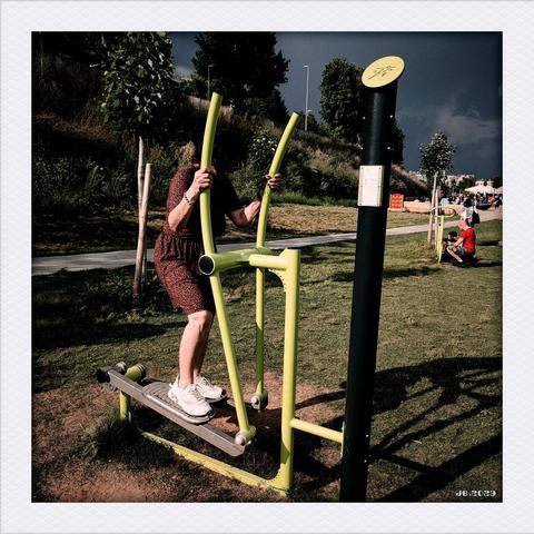 Farbfoto, das eine Frau in einem braunen Kleid und weißen Sportschuhen auf einem Crosstrainer im Freien in einem Park zeigt. Weiter entfernt ist ein zweites Sportgerät mit einer weiteren Person zu sehen.