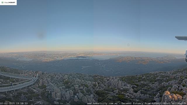 A panoramic view from Kunanyi/Mount Wellington, showcasing the Derwent Estuary and surrounding landscape. The scene includes rocky terrain in the foreground, with a winding path and low vegetation, and a distant view of the water and hills under a clear sky. I’m standing on a walkway on the left looking up at the camera.