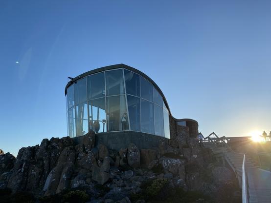 A modern, glass-encased structure sits on rocky terrain, surrounded by nature, atop Kunanyi. The building has a curved roof and offers viewing points. It is illuminated by the setting sun, with people visible near the entrance and on a wooden path leading to it