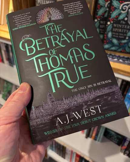 A copy of the beautiful novel, The Betrayal of Thomas True, being held up against a background of filled bookshelves.