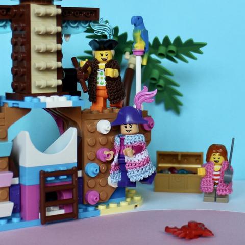 3 Lego pirate minifigs climbing a playground pirateship decorated with blue, pink and white parts.
In the back a palm tree and a gold treasure chest.
