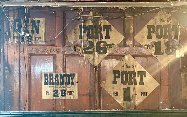 Wood-panelled wall with five letterpress printed posters pasted to it. They are old and well-worn, and advertise the prices of gin, brandy and port (x3). Across the middle portion of the wall is signwritten lettering that isn't completely visible, as per the text in the main post.