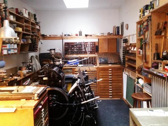 Blick in die Druckerei: Ein schmaler Raum mit Oberlicht, in der Mitte steht eine große Druckmaschine, der Heidelberger Tiegel. Umlaufend sind Regale und Schränke mit Werkzeugen, Farbdosen und Beischrift.