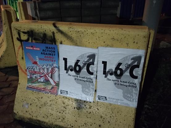 Een betonblok met daarop enkele affiches.
Op de Code Rood affiche staat "Mass action against agro-industry - 28/02 - 02/03 Belgium" en een gekruiste vork met riek.
Op de andere affiche staat "1.6°C - The earth is not dying, it is being killed" en een schedel op de achtergrond.