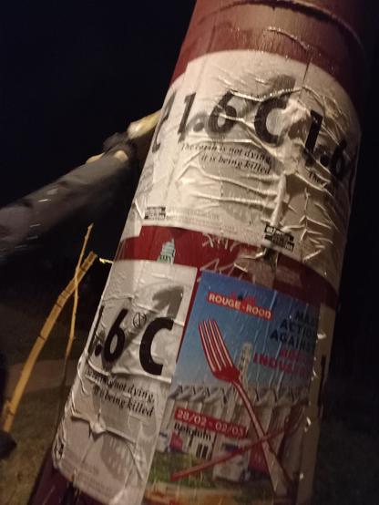 Een pillaar met verschillende affiches op geplakt.
Op de Code Rood affiche staat "Mass action against agro-industry - 28/02 - 02/03 Belgium" en een gekruiste vork met riek.
Op de andere affiche staat "1.6°C - The earth is not dying, it is being killed" en een schedel op de achtergrond.