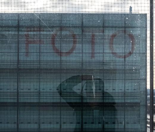 Das Bild zeigt eine komplexe Komposition aus Reflexionen, Transparenz und strukturellen Details. Im Vordergrund befindet sich ein mit einem Schutznetz bedecktes Fenster, das sowohl den Blick nach draußen als auch die Reflexion eines Fotografen zeigt. Der Fotograf, dessen Silhouette und Haltung sichtbar sind, scheint die Szene aufzunehmen, was eine zusätzliche Dimension des Selbstbezugs einbringt.

Hinter dem Netz und der Scheibe ist ein Gebäude zu sehen, auf dessen Glasfassade in großen roten Buchstaben das Wort „FOTO“ geschrieben steht. Die Buchstaben sind leicht verblasst, was ihnen einen gealterten oder subtilen Charakter verleiht. Das Netz und die Struktur des Fensters fügen eine geometrische Textur hinzu, die die visuelle Tiefe und Vielschichtigkeit des Bildes verstärkt.

Im Hintergrund ist ein heller Himmel mit leichter Bewölkung zu erkennen, der das Licht gleichmäßig über die Szene verteilt. Die kühlen Farbtöne dominieren das Bild, was eine ruhige, fast introspektive Stimmung vermittelt.

Die Komposition wirkt durch die Überlagerung von Innen- und Außenwelt, durch Text, Reflexion und Perspektive kunstvoll und nachdenklich. Das Bild thematisiert möglicherweise die Beziehung zwischen dem Fotografen, der Umgebung und der Wahrnehmung selbst.