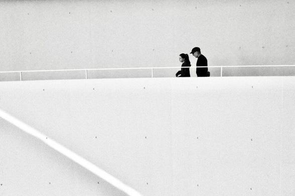 Das Bild zeigt eine minimalistische Szene in Schwarz-Weiß. Zwei Personen gehen nebeneinander entlang einer oberen Plattform oder Galerie, die durch ein schlichtes Geländer abgegrenzt ist. Die Personen tragen dunkle Kleidung und heben sich stark von der hellen, fast leeren Umgebung ab. Eine der Personen trägt eine Mütze, während die andere einen Pferdeschwanz hat. Sie scheinen ruhig zu sein, möglicherweise in ein Gespräch vertieft oder in Gedanken versunken.  Die Umgebung ist modern und reduziert, mit klaren Linien und einer dominanten weißen Fläche im Vordergrund, die einen großen Teil des Bildes einnimmt. Diese Fläche könnte eine Wand oder eine geneigte Ebene sein und verleiht dem Bild eine abstrakte und geometrische Ästhetik. Kleine Details, wie die sichtbaren Schrauben oder Befestigungen auf der Fläche, betonen die Kargheit und den architektonischen Charakter der Szene.  Das Bild vermittelt eine ruhige, introspektive Atmosphäre und spielt mit Kontrasten zwischen den dunklen Silhouetten der Personen und der minimalistischen, hellen Umgebung. Die Komposition wirkt bewusst zurückhaltend und lässt Raum für Interpretation und emotionale Resonanz.