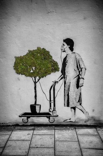 A powerful street art piece by Dr. Love depicting a woman in a hospital gown, barefoot, using an oxygen mask connected to a moss-covered structure shaped like a small tree instead of an oxygen tank. The moss forms the tree’s foliage, creating the illusion of a living plant placed on a wheeled cart as if it were a life-support machine. The mural is painted in black and white, contrasting with the vibrant green of the moss, emphasizing themes of environmental dependence and the necessity of nature for survival. The artwork is located on a textured concrete wall, blending stencil work with real-life elements for a striking visual impact.