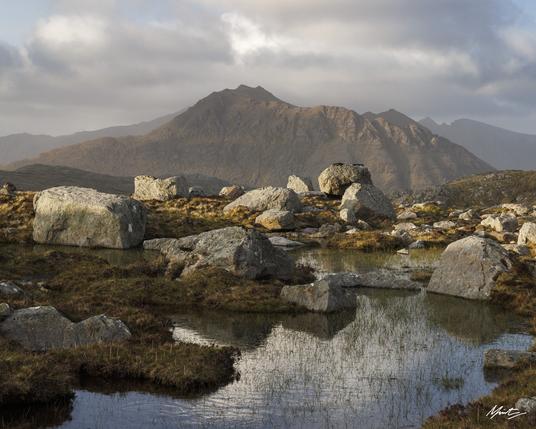 a small lochan high in the remote mountains of scotland reflects beautiful clouds and blue skies. erratic boulders are strewn across the foreground while the background features a shapely mountain with a steep and wandering ridgeline. soft evening light dances across the scene.
