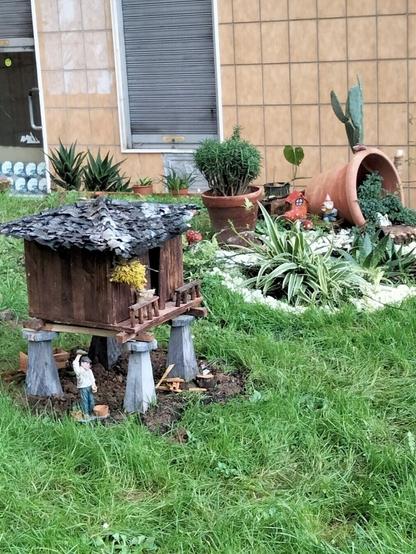 En un pequeño jardín, un hórreo en miniatura cobija a un paisano echando sidra. El hórreo esta hecho de madera marrón, con el tejado de pizarra y los pegoyos simulando piedra. Al fondo se ven macetas de diferentes tamaños con plantas. Al fondo a la derecha, un parterre de piedra blanca rodea plantas de hoja. Una gran maceta está tumbada, brotando otra planta de ella.