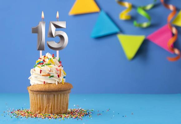 Ein mit weißem Zuckerguss und bunten Streuseln überzogener Cupcake mit Metallkerzen in Form der Zahl 15. Der Hintergrund besteht aus lebhaften dekorativen Elementen wie dreieckigen Bannern und Luftschlangen, die sich vor einem blauen Hintergrund abzeichnen.