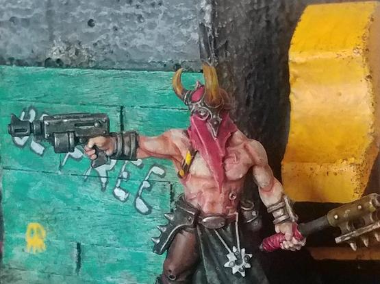 A bare chested man aims his pistol. His skin is pale, and he has a horned red hood covering his head. He also carries a spiked hammer. A white star dangles from his belt. He stands before a concrete wall, and to his right are teal boards with graffiti, while his left is in front of a big chunk of metal, striped yellow and black.