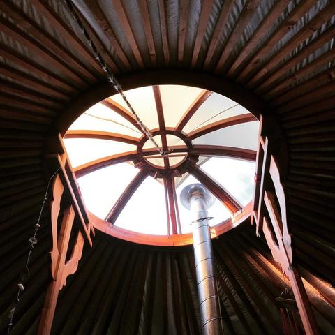 🇩🇪 Innenansicht des kreisförmigen Oberlichts einer Jurte, die einen Holzrahmen und ein vom Boden aufsteigendes Metallofenrohr zeigt. An den Seiten sind Holzstützen sichtbar, an denen kleine Zierelemente hängen. Das Oberlicht lässt helles Licht herein. 
🇬🇧 Interior view of a yurt's circular skylight, showing a wooden frame and a metal stovepipe rising from the floor. Wooden supports are visible along the sides, with small decorative elements hanging from one. The skylight lets in bright light.