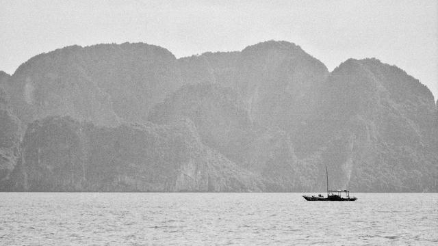 A series of black and white photographs capturing traditional wooden boats navigating calm waters, set against dramatic limestone karst formations. The images depict solitary and grouped boats floating in misty bays, with towering cliffs, distant pagodas, and small lighthouses adding depth to the serene landscapes.