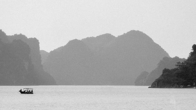 A series of black and white photographs capturing traditional wooden boats navigating calm waters, set against dramatic limestone karst formations. The images depict solitary and grouped boats floating in misty bays, with towering cliffs, distant pagodas, and small lighthouses adding depth to the serene landscapes.