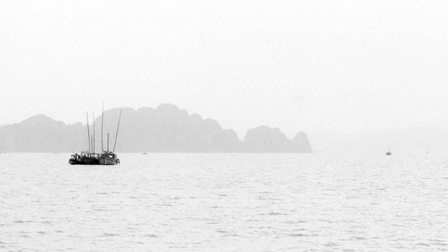 A series of black and white photographs capturing traditional wooden boats navigating calm waters, set against dramatic limestone karst formations. The images depict solitary and grouped boats floating in misty bays, with towering cliffs, distant pagodas, and small lighthouses adding depth to the serene landscapes.
