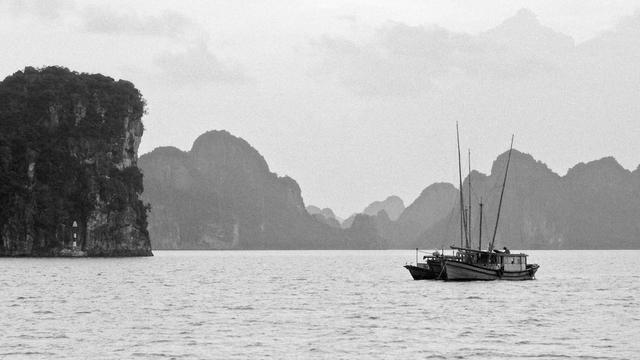 A series of black and white photographs capturing traditional wooden boats navigating calm waters, set against dramatic limestone karst formations. The images depict solitary and grouped boats floating in misty bays, with towering cliffs, distant pagodas, and small lighthouses adding depth to the serene landscapes.