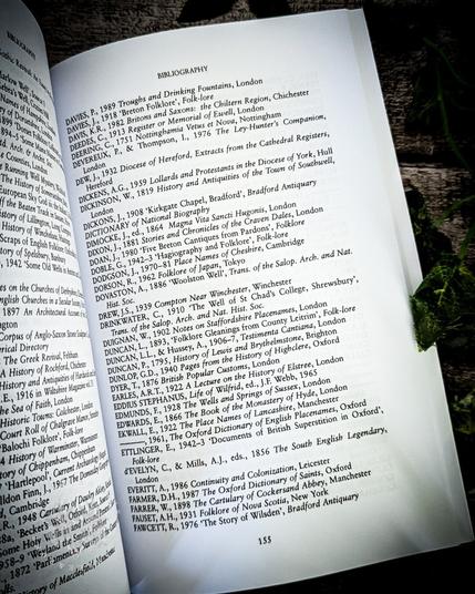 A close-up image of a page from the book "The Living Stream: Holy Wells in Historical Context." The page shows the bibliography section, with several references listed. Some titles include "Troughs and Drinking Fountains," "Britons Folklore," and "Documents of British Superstition in Oxford." The page is surrounded by green leaves, and the text is clearly visible, with a focus on the academic references related to holy wells and folklore studies. The book’s content is scholarly and detailed.