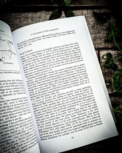 A page from The Living Stream: Holy Wells in Historical Context titled "At the Heart of the Community." This section explores the social control associated with holy wells, particularly in relation to the English and Celtic experiences. It discusses how the Catholic religious practices at these sites changed post-Reformation, with some local rituals surviving through adaptations. The text highlights the social and political functions of holy wells and their connection to both the church and the community. The page is photographed on a rustic wooden surface with green ivy leaves.