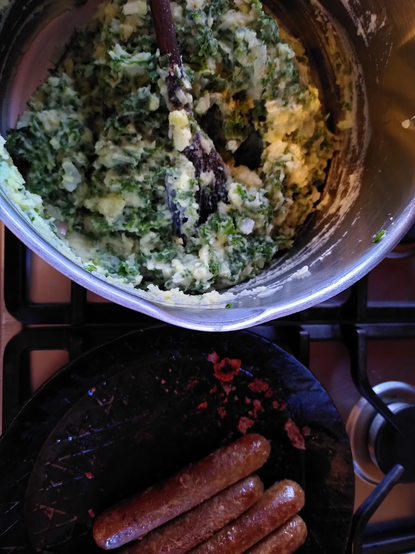 Potkieker: Du guckst von oben in einen großen Kochtopf mit Colcannon und eine Bratpfanne mit vier angebratenen groben Bratwürsten. Topf und Pfanne stehen auf einem Gasherd.