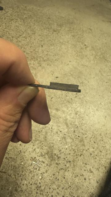Eine Person hält ein kleines, flaches Metallstück mit einer Kerbe auf einer Seite gegen eine strukturierte Betonoberfläche. Das Metallstück ist ein Bremsbelag einer Scheibenbremse. Der Belag ist noch einwandfrei.