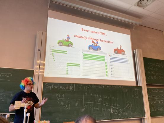 Robert wearing a clown wig, gesturing hands and looking serious, in front of a chalkboard and projector screen in a class room at University Libre Brussels at the web performance track at Fosdem 2025.