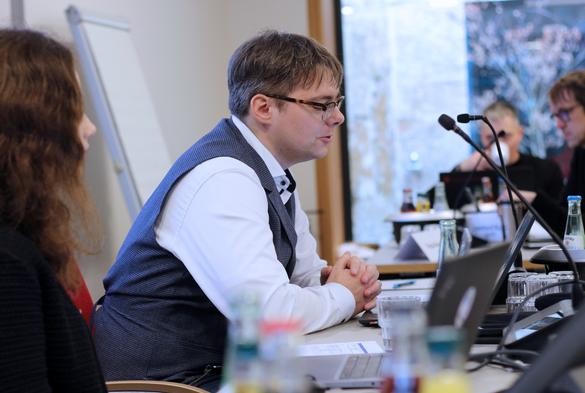 A man in a white shirt and vest speaking into a microphone during a meeting or conference. He is seated at a table with laptops, documents, and water bottles. Other attendees are visible in the background.