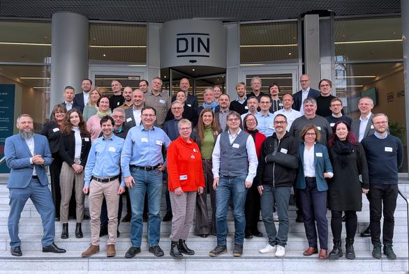 A large group of people posing for a photo on the steps outside a building with a "DIN" sign above the entrance. The attendees are wearing name tags and a mix of formal and casual clothing, smiling at the camera.