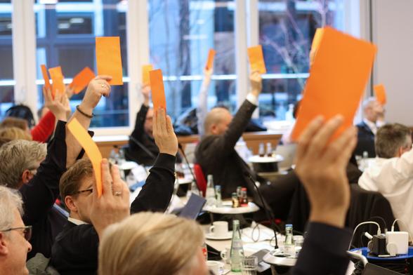 A group of people in a meeting or conference room raising orange cards in a voting process. The room has tables with water bottles, coffee cups, and documents, and large windows showing an outdoor view.