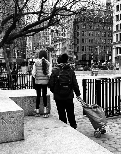 A black-and-white photo of the back of a woman and young girl standing in Madison Square, Park and staring up at the flat iron building. The young girl is on a pedestal and wearing silver rollerskates. There are gates around the park and many city buildings can be seen in the background