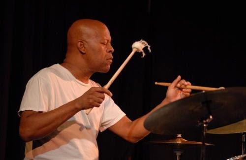 William Hooker seated at his drum kit. His eyes are closed as he plays, suggesting deep concentration.