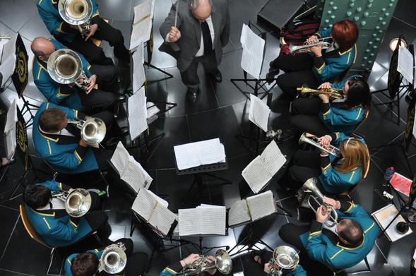 The image shows a top-down view of a musical ensemble, likely a brass band, with musicians seated in a circular arrangement. They are playing instruments such as cornets, euphoniums, and tubas. A conductor stands in the center with a baton, leading the group. Sheet music is placed on stands in front of each musician. The members appear to be wearing matching uniforms.