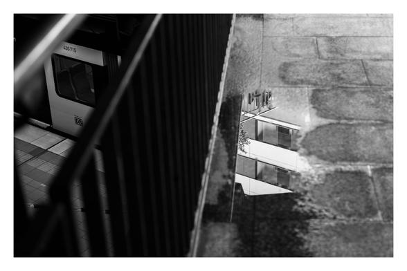 Schwarz-weiß Foto im Querformat. Blick Richtung Boden an einem Bahnhof. Mittig geht ein Geländer von oben nach unten durchs Bild. Rechts davon ist am Boden eine Pfütze, in der sich die Fenster eines Hauses spiegeln. Links vom Geländer geht der Blick ein Stockwerk tiefer aufs Gleis. Ein kleiner Ausschnitt der S-Bahn ist zu sehen. Auf den ersten Blick sieht es so aus, als ob sich die Bahn in der Pfütze spiegeln würde bzw. die Spiegelung in der Pfütze die Fortsetzung des Zuges ist.
