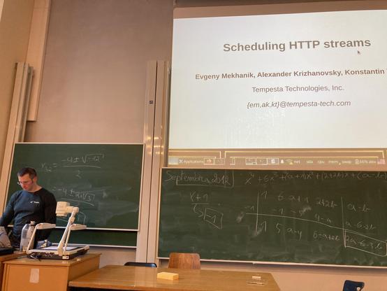 Alexander presenting on stage at the Fosdem Web Perf track in a lecture room at University Libre Brussels, next to a his slides and a green chalkboard.
