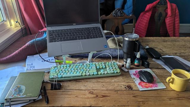 A table with laptop, keyboard, mouse, journals, coffee, yeti water mug, kindle, book