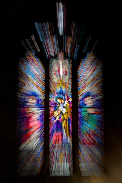 A stained glass window made up of three main panels, narrow and tall with smaller sections at the top making an arch at the top of the window. However, none of this is too clear because the light from the window has a motion blur, making it look like coloured light beams shining in through the window. The three main panels depict people, but no real detail can be made out.